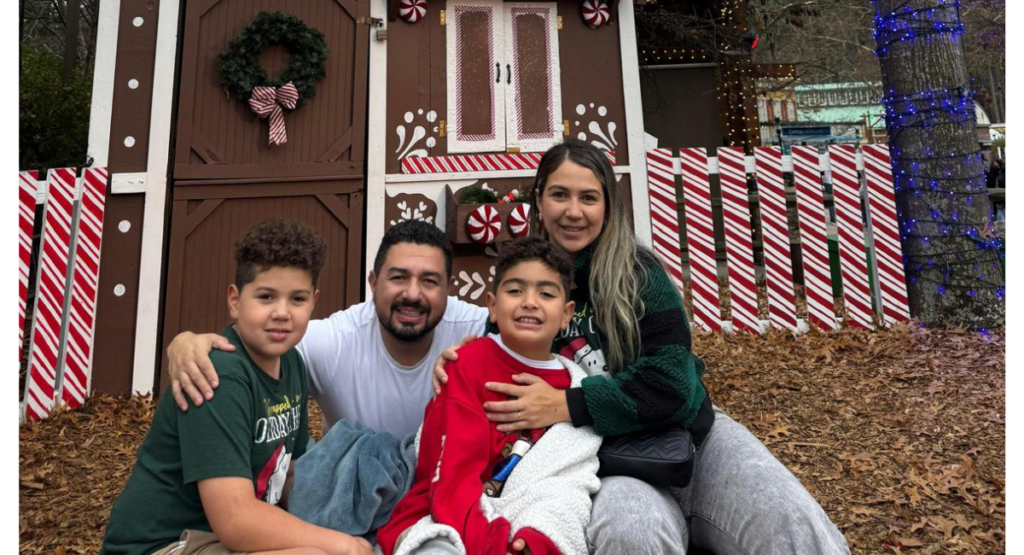 Cesar, his wife, and his two sons sit on the ground outdoors in front of a Christmas holiday scene.