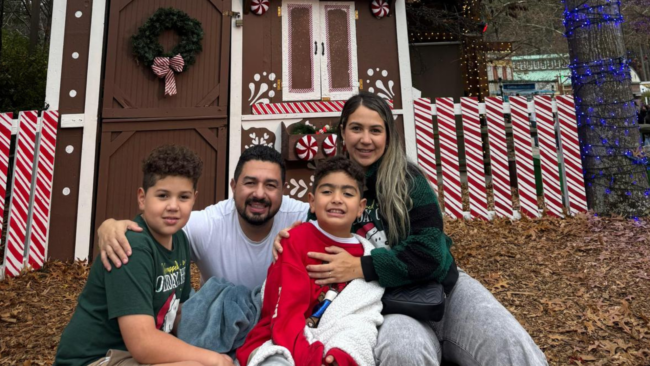 Cesar and his family pose outdoors in front of a large gingerbread house.